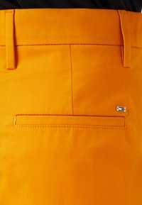 Oranžové látkové kalhoty s zadní kapsou a čistým šitím, na které je malý kontrastní akcent loga. Hladká textura a přizpůsobený design.