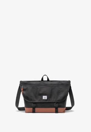 Messenger bag with a black canvas body, brown bottom, adjustable strap, and front flap with a logo patch and zippered pocket.
