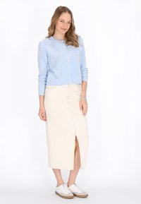 Vaalean sininen neulecardigan, jossa on tekstuureilla varustettu kuvio, yhdistettynä kermaiseen midihameeseen, jossa on etuhalkio, ja valkoisiin lenkkareihin.