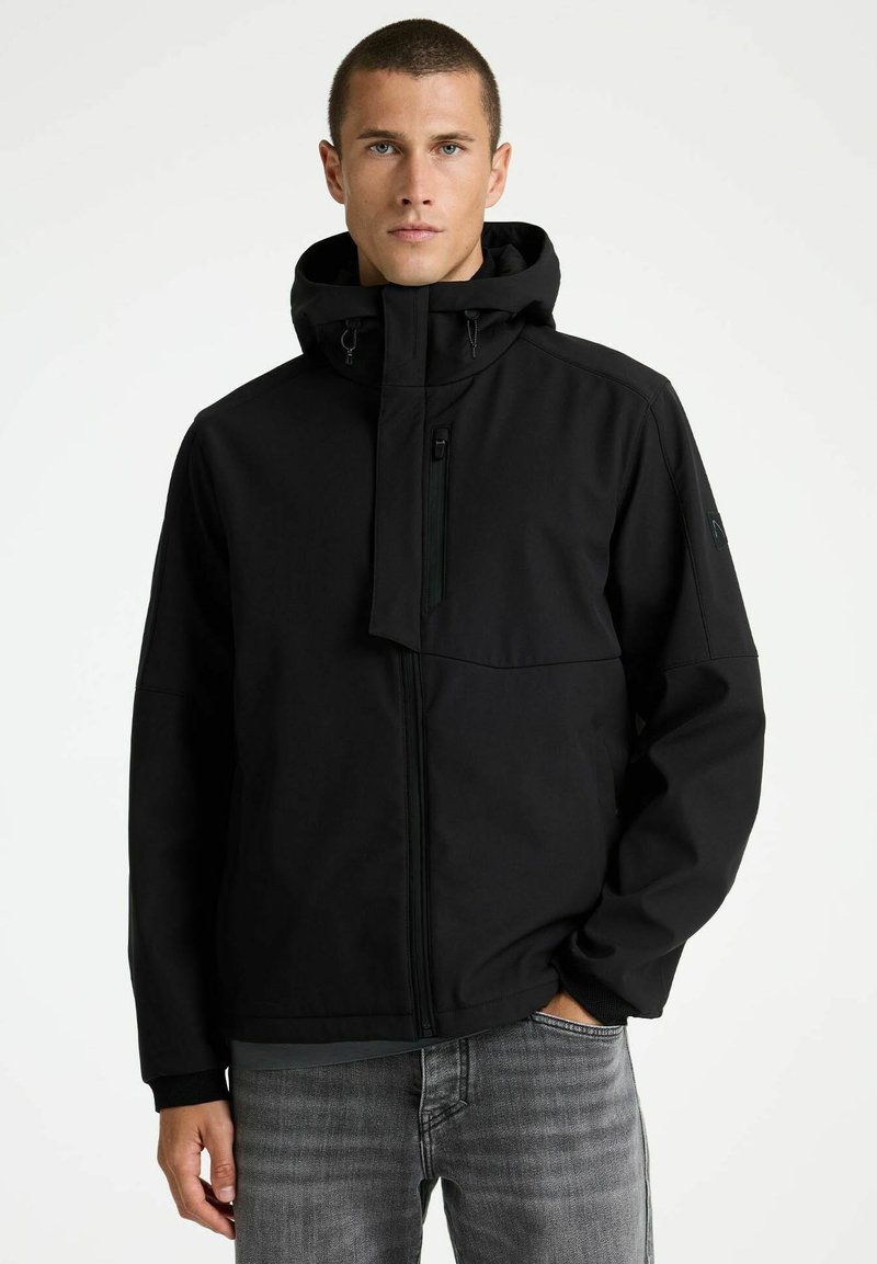 Mees, kes kannab musta kapuutsiga jope, millel on lukk ja rinnatasku, seisab ühe käega hallide teksataskus valgel taustal.