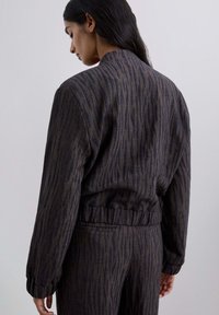 Chaqueta de patrón oscuro con una superficie texturizada, que presenta una cintura elástica y puños acanalados, mostrada desde un ligero ángulo en la parte posterior.