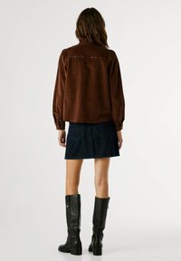 Camisa de ante marrón de manga larga con puños abotonados, combinada con una mini falda de mezclilla oscura y botas de cuero negro hasta la rodilla.