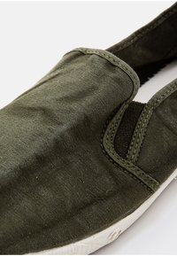 Zöld vászon slip-on cipő, texturált felülettel, rugalmas oldalakkal és fehér gumi talppal, varrott részletekkel.