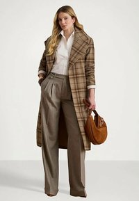 Γυναίκα που φοράει ένα καφέ καρό μακρύ παλτό, άσπρη μπλούζα, γκρίζες παντελόνας σε φαρδιά γραμμή, κρατώντας μία καφέ σουέτ τσάντα.