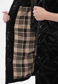 Puchowa czarna odzież wierzchnia o błyszczącej powierzchni, z beżowym i czarnym kratkowanym podszewkiem. Zapinana na guziki wzdłuż krawędzi.