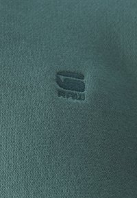 Gros plan sur un tissu tricoté bleu sarcelle avec un logo brodé bleu sarcelle foncé présentant un "G" stylisé et le texte "RAW" en dessous.