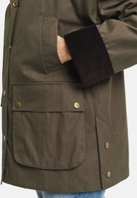 Olive green outerwear with a corduroy cuff, featuring two front pockets and brass snap buttons. Smooth texture with a relaxed fit design.