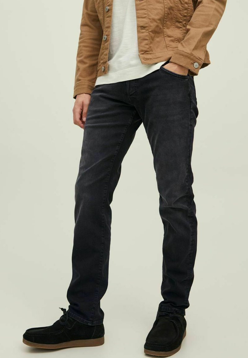 Jeans oscuros desgastados con un corte ajustado, combinados con una camisa beige clara y una chaqueta marrón, complementados con zapatos de ante negros.
