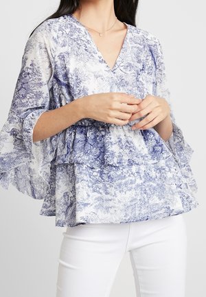 Kvinde iført en blå og hvid blomsterprintet V-hals bluse med lagdelte flæseærmer og hvide bukser, hænderne foldet ved taljen.