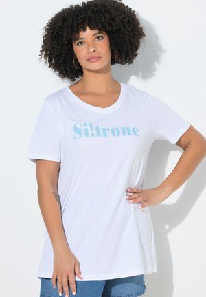 Frau mit lockigem Haar, die ein weißes „Si!trone“ T-Shirt und Jeansshorts trägt, steht mit einer Hand in der Hüfte vor weißem Hintergrund.