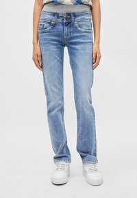 Ανοιχτού μπλε τζιν με εφαρμοστή μέση, κλείσιμο με δύο κουμπιά, μπροστινές τσέπες και διακριτές ραφές. Συνδυασμένα με λευκά αθλητικά παπούτσια.