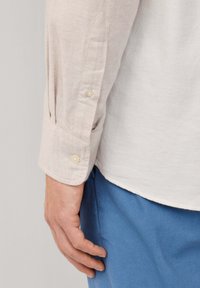 Lichtbeige overhemd met lange mouwen en opvallende manchetten met twee knopen, gemaakt van een gestructureerde stof. Blauwe broek is zichtbaar aan de onderkant.