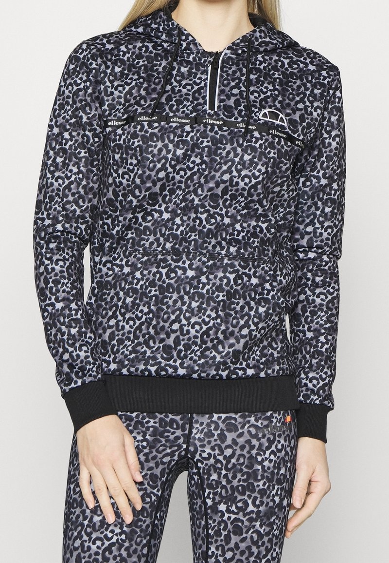 Femme portant un sweat à capuche et des leggings assortis imprimés léopard noir et gris, avec des poignets noirs et un logo blanc de la marque sur la poitrine.