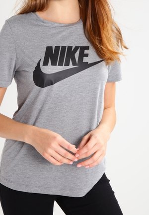 Nainen yllä harmaa Nike-t-paita, jossa musta logo, kädet ristissä edessä, näkyvissä ylävartalo ja osa mustista housuista.