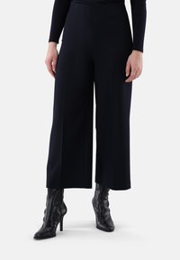 Weit geschnittene Hose aus dunkelblauem Stoff mit glatter Textur, Seitentaschen und vorderen Falten, kombiniert mit eleganten schwarzen Stiefeletten.