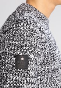 Maglione lavorato a maglia in bianco e nero con un motivo testurizzato. Presenta un collo rotondo e una toppa in pelle nera con logo in rilievo.