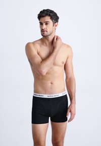 Sorte bomullsboxer med en hvit elastisk midjebånd som har "BJÖRN BORG"-logo, modellen står mot en ensfarget bakgrunn.