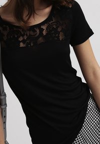 Kvinna i svart kortärmad topp med blommig spetsplattform och byxor med houndstooth-mönster, håller en ljusgrå väska.