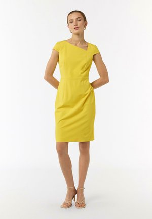 Gelbes, tailliertes Kleid mit kurzen Ärmeln, schrägem Ausschnitt und definiertem Bund. Glattes Material mit einem knielangen Saum, kombiniert mit beigen High-Heels.