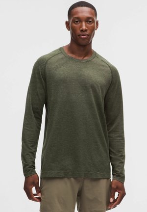 Ung mand iført en olivengrøn tekstureret langærmet skjorte og lysebrune shorts, der står mod en ensfarvet baggrund.