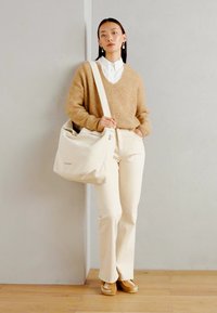 Beige tote bag laget av mykt materiale, med justerbar stropp og subtil logo, kombinert med lyse bukser og en strikket genser.