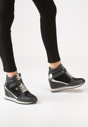 Personne portant des baskets compensées noires avec des lacets blancs et des accents gris, associées à un pantalon noir ajusté sur un fond uni.