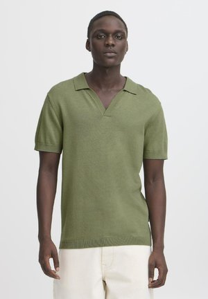 Short-sleeve, V-neck shirt in olive green knit fabric. Features a relaxed fit and ribbed hem. Worn with light-colored trousers.
