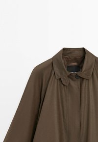 Brun langærmet frakke med en bred krave og en enkelt synlig sort knaplukning ved halsen.