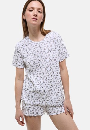 Femme portant un haut blanc à manches courtes et un short avec un petit motif floral, debout devant un fond clair uni.