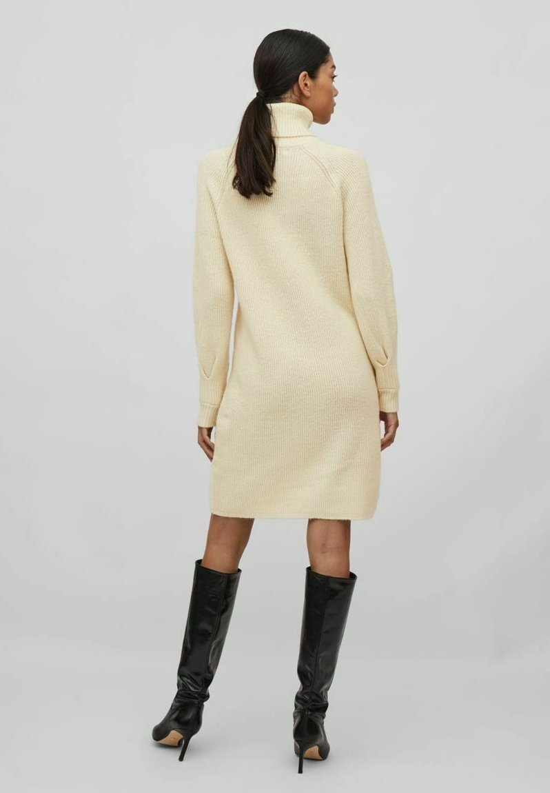 Robe en maille à col roulé de couleur crème claire, avec une texture côtelée et des manches longues. Associée à des bottes noires brillantes montantes.