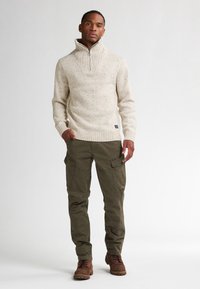 Maglione beige in lana con colletto alto e texture a coste, abbinato a pantaloni cargo verde oliva dotati di tasche laterali, indossati con stivali marroni.