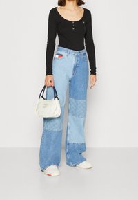 Schwarzer langärmeliger Bodysuit mit Knöpfen, kombiniert mit blauen Patchwork-Weitbeinhosen. Hält eine kleine weiße Handtasche mit einem marineblauen Griff.