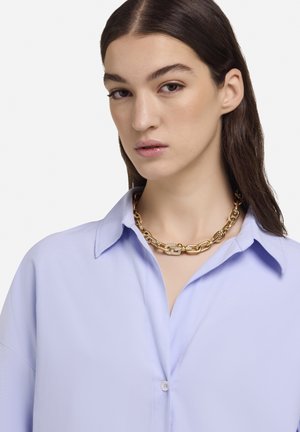 Gouden ketting met gestructureerde schakels en een centrale versiering, gedragen over een lichtblauw overhemd met knopen en een kraag.