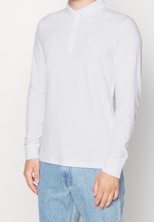 Polo-shirt blanc à manches longues avec un col et une patte de boutonnage à trois boutons. Doté d'un logo texturé sur la poitrine et de poignets ajustés. Porté avec un jean bleu.