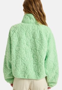 Green fleece jacket featuring a textured floral pattern, high collar, and relaxed fit. The fabric appears soft and thick.