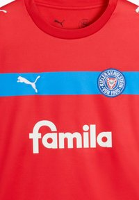 Maillot rouge avec une bande horizontale bleue et le texte "famila" en blanc. Présente un écusson circulaire rouge avec un design de bouclier et un logo Puma blanc.