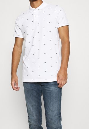 Homme portant un polo blanc à manches courtes avec de petits motifs d'oiseaux noirs et un jean bleu, debout devant un fond uni.