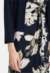 Marineblauwe cardigan over een bloemenjurk met crème- en olijfgroene patronen, met een geknoopte details bij de taille. Zachte stoftextuur.