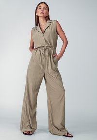 Combinaison sans manches en tissu beige clair avec un col, des boutons à l'avant, une taille élastique et des jambes larges, dotée de poches latérales.