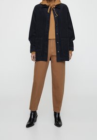 Chaqueta de pana oscura con cierre de botones y dos bolsillos frontales, superpuesta a una sudadera marrón, combinada con pantalones beigest y botas de tacón negras.