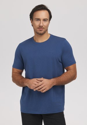 Mężczyzna o krótkich ciemnych włosach, ubrany w niebieski krótki t-shirt i czarne spodnie, stoi ze złożonymi rękami z przodu.