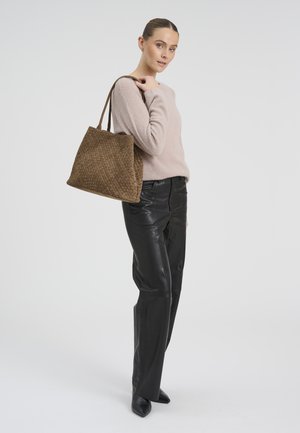 Naine, kes kannab beežikat kampsunit ja musti nahkpükse, hoides õlal suurt pruuni punutud käekotti, seisab ühevärvilise tausta ees.