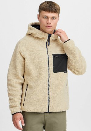 Beige fleece jacket with a hood, featuring a black zippered chest pocket, side zipper pockets, and ribbed cuffs.