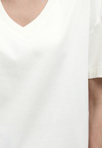 Κοντινό πλάνο ενός ατόμου που φοράει απλό λευκό κοντομάνικο μπλουζάκι με V λαιμόκοψη, με ορατό το πάνω μέρος του στήθους και μέρος του χεριού.