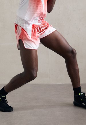 Legs of a person wearing red and white boxing shorts and black Venum boxing shoes, positioned in a boxing stance on a gray floor.