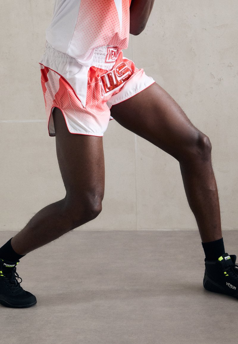 Legs of a person wearing red and white boxing shorts and black Venum boxing shoes, positioned in a boxing stance on a gray floor.