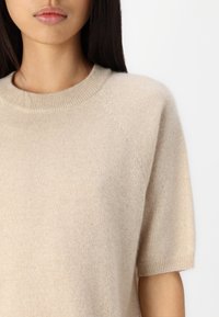 Beżowy sweter dziany z krótkim rękawem, żebrowanym dekoltem i miękkim, teksturowanym wykończeniem. Casualowy, luźny krój z raglanowymi szwami.
