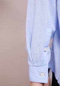 Světle modrý rukáv dlouhého trička s knoflíkovým zapínáním, viditelná ruka a neutrální béžové pozadí.