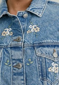 Chaqueta de denim con un lavado azul claro que presenta bordados florales en blanco y melocotón. Incluye botones plateados y un diseño de cuello clásico.