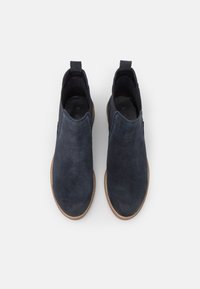 Un paio di stivali alla caviglia in suede blu scuro, con suole color terra, pannelli elastici laterali e linguette, visti dall'alto su uno sfondo bianco.